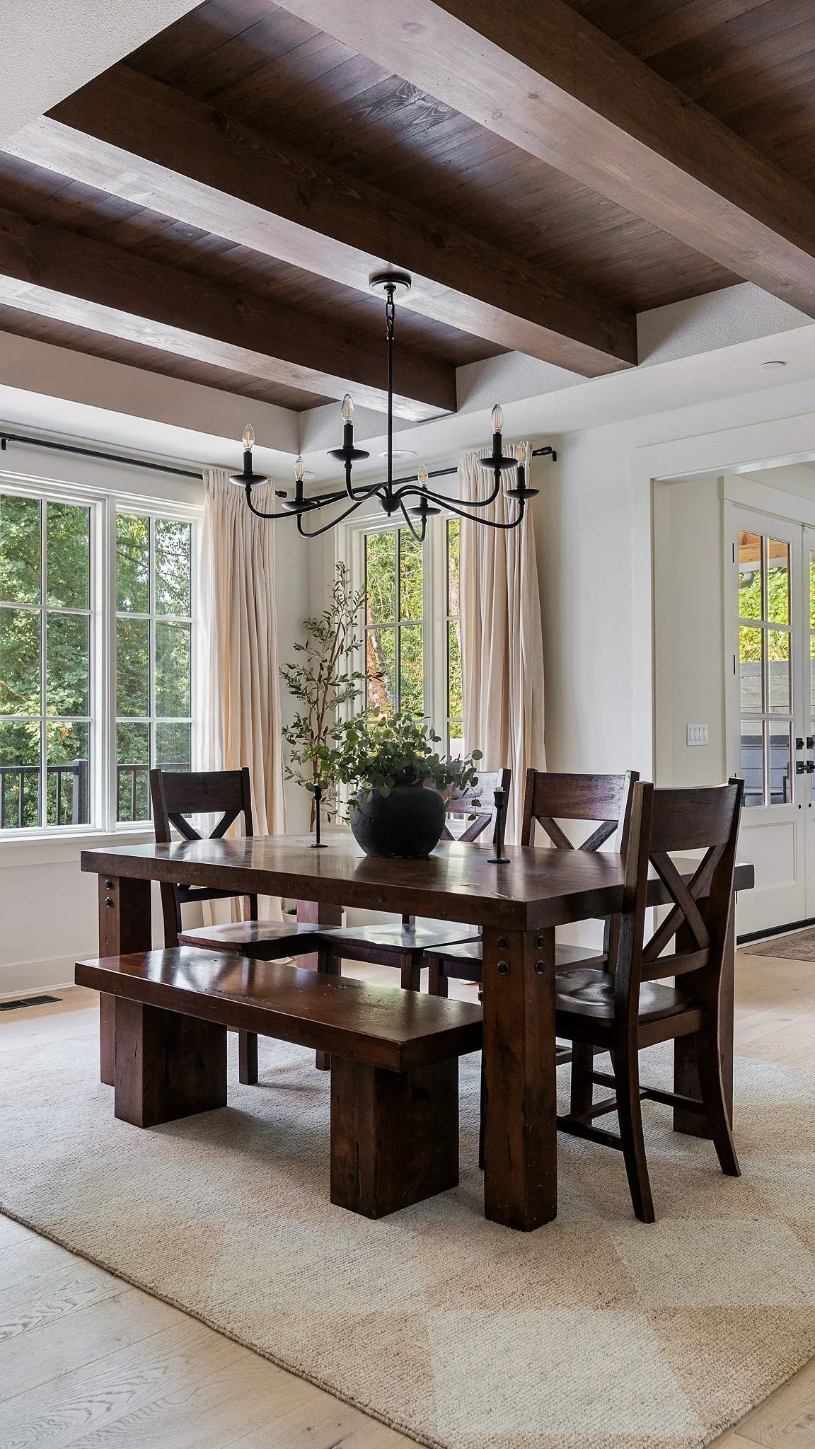 Dining room featuring a rustic dark wood table, bench, and chairs illuminated by a black metal chandelier beneath wood ceiling beams.