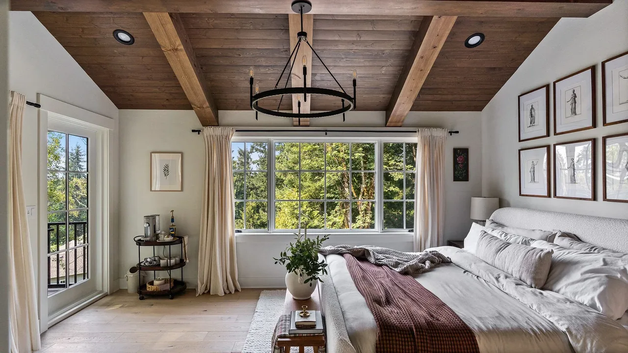 A bedroom featuring a wood beam ceiling, large multi-pane window with beige curtains, and white bedding with a burgundy throw at Cougar Mt Custom by H2D architecture + design.