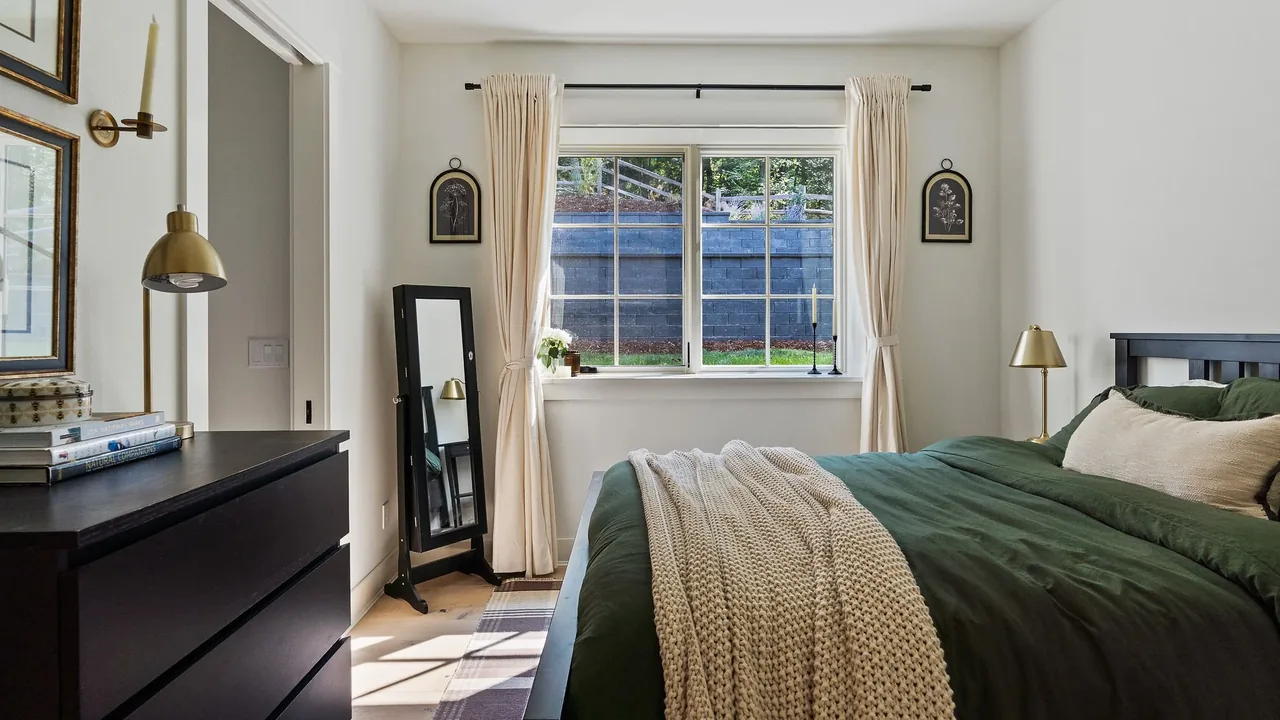 A bedroom featuring a dark-framed bed with green bedding, cream curtains, and a standing mirror beside the window.