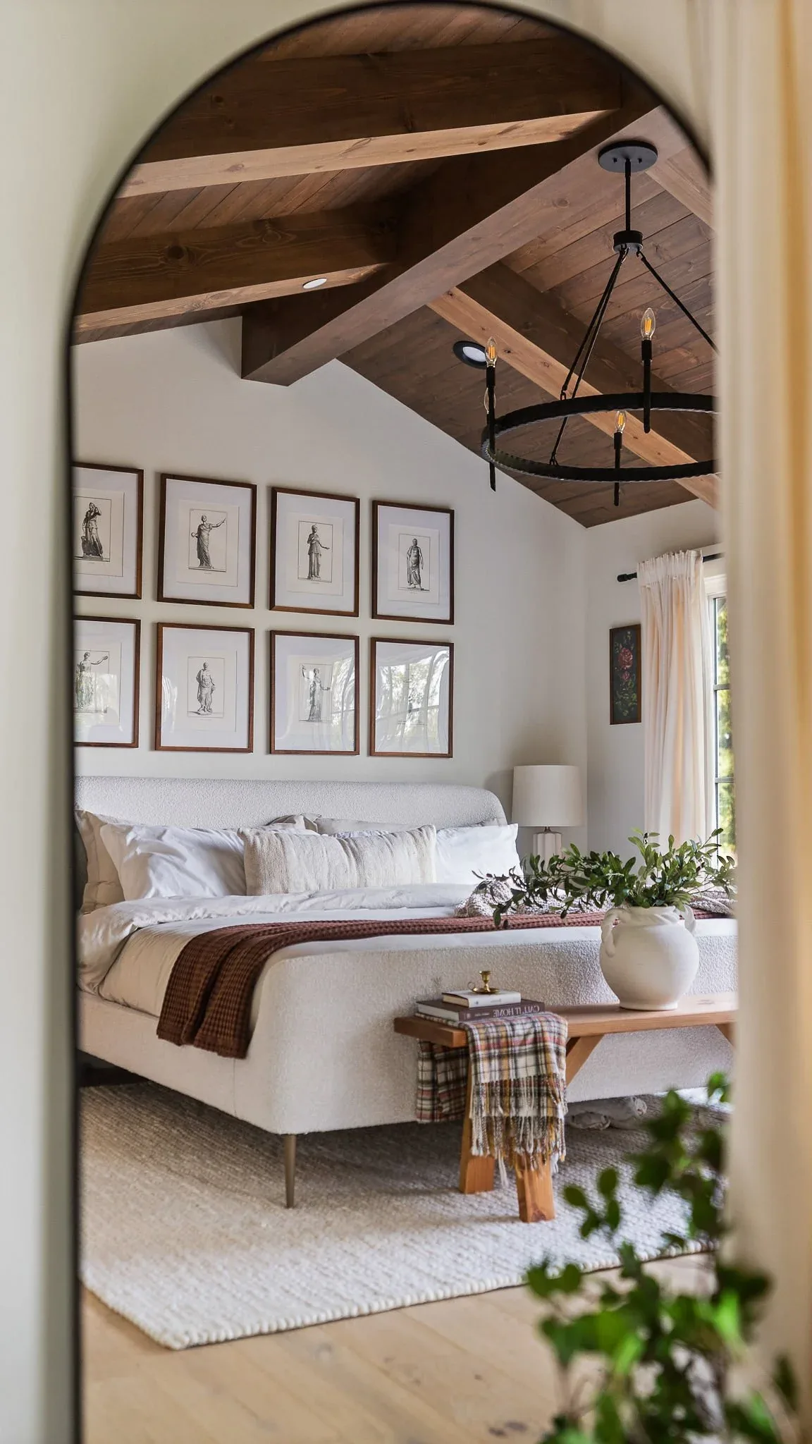 Bedroom with vaulted wood ceiling, exposed beams, white upholstered bed, and black metal chandelier.