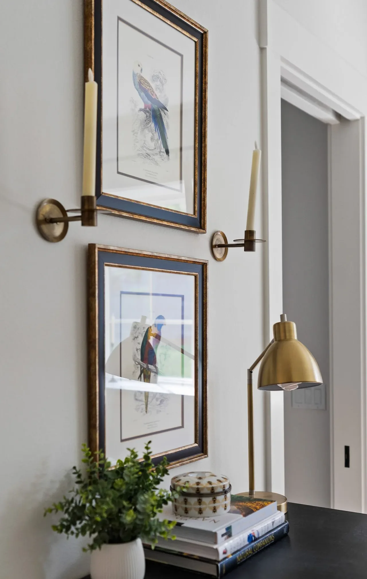 Bird-themed artwork framed on a wall flanked by brass candle sconces with a gold desk lamp on a nearby surface.