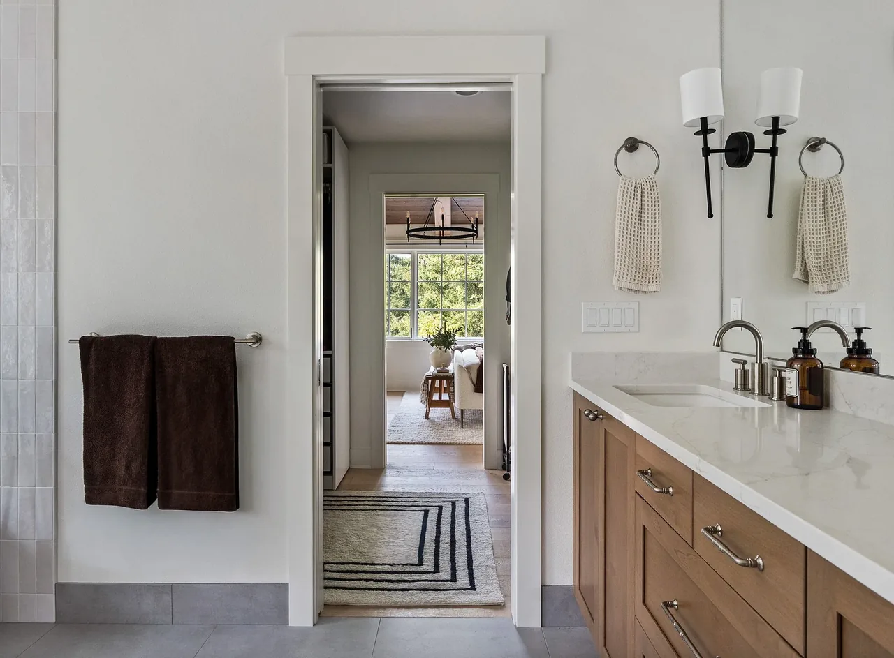 Bathroom with wood cabinetry vanity and metal hardware opens toward a living area with sofa and large window.