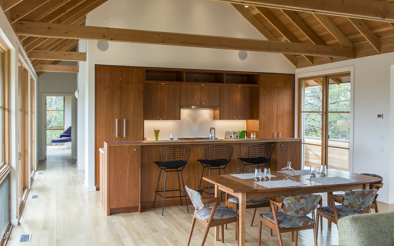 Kitchen and dining area with wooden ceiling beams and cabinetry, featuring a kitchen island with black metal bar stools.