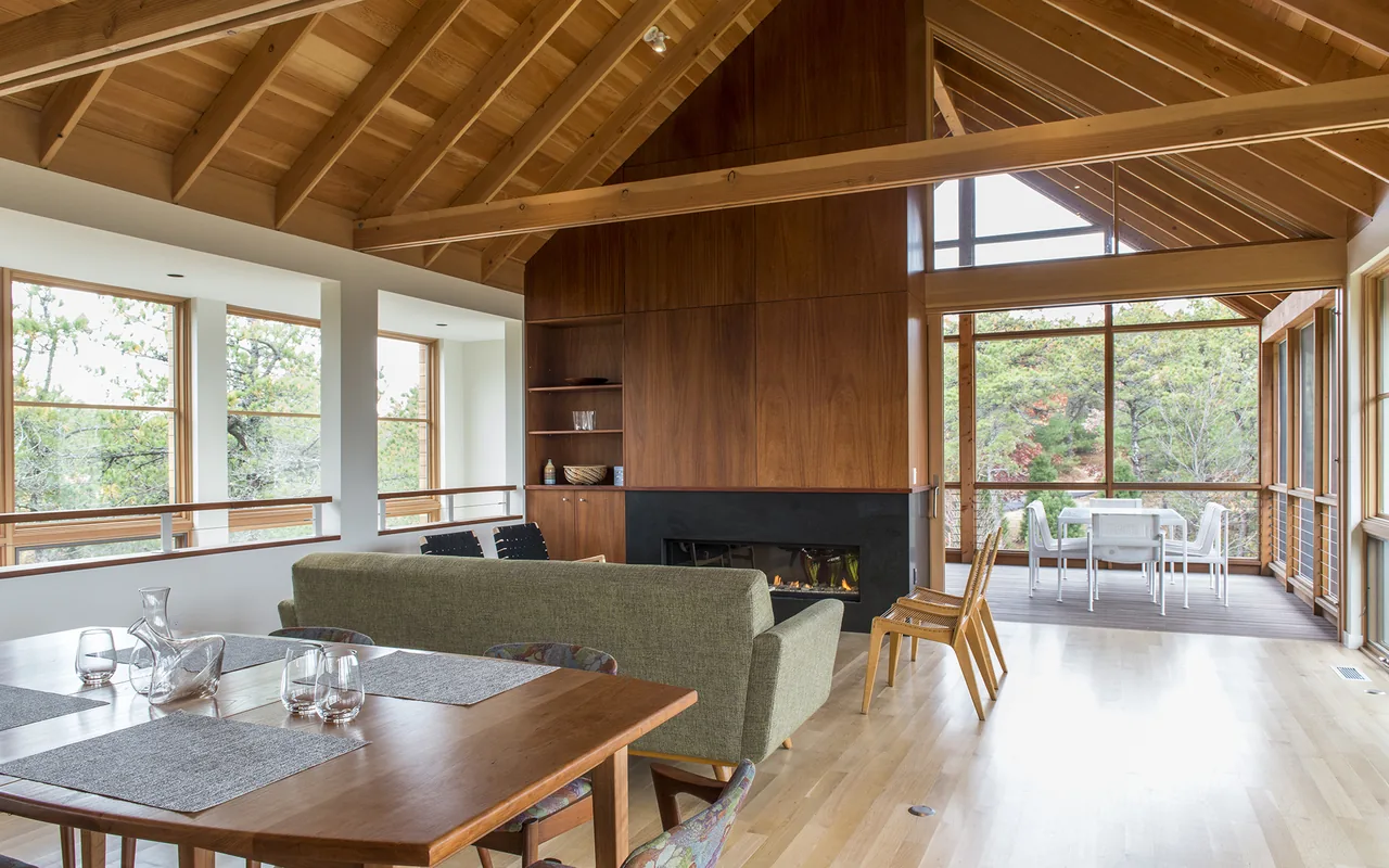 Living and dining area with vaulted ceiling, wood panel fireplace, and large windows overlooking trees.