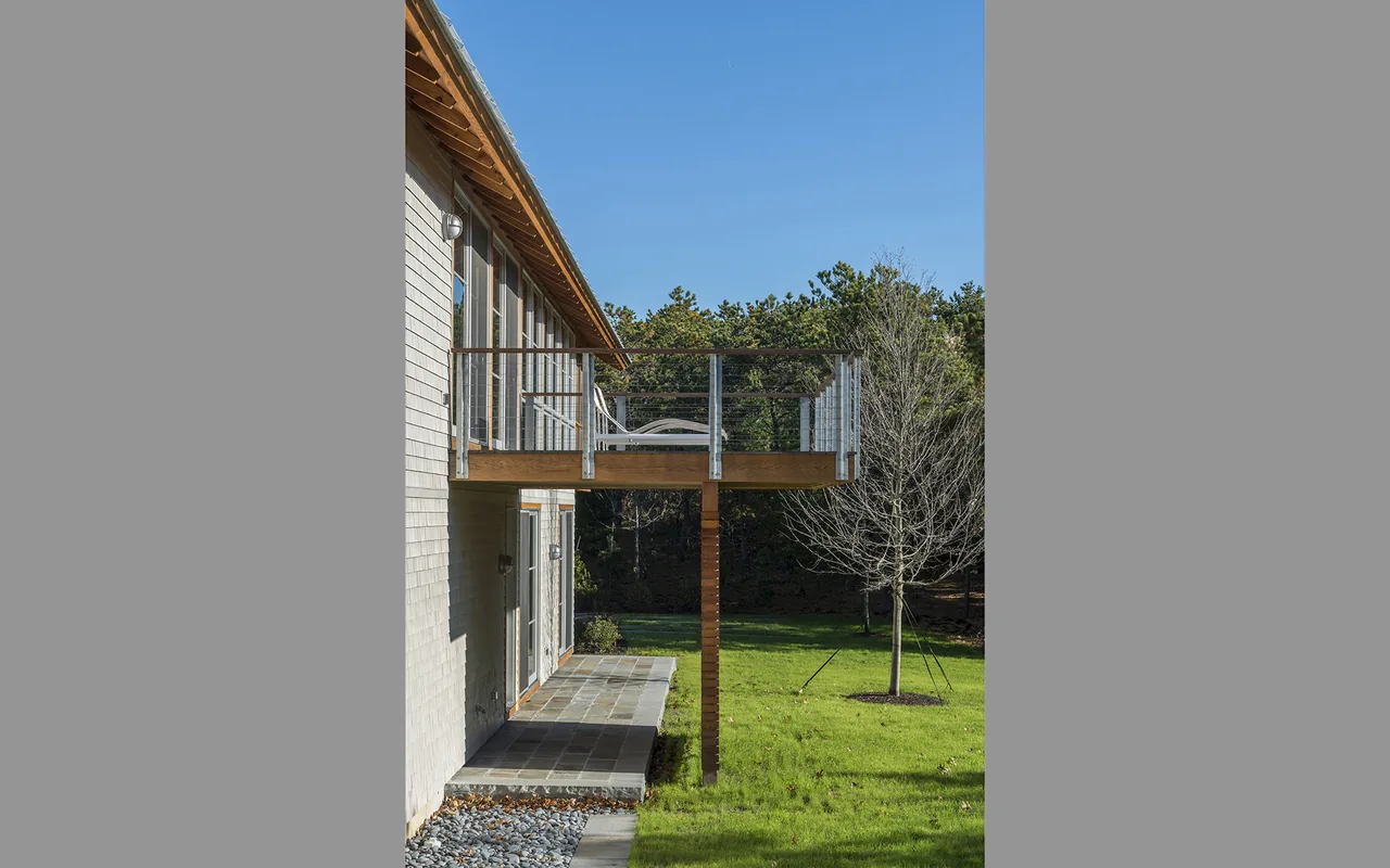 The north-side elevation shows a cantilevered balcony with glass railing and a white outdoor chair overlooking the grassy backyard.