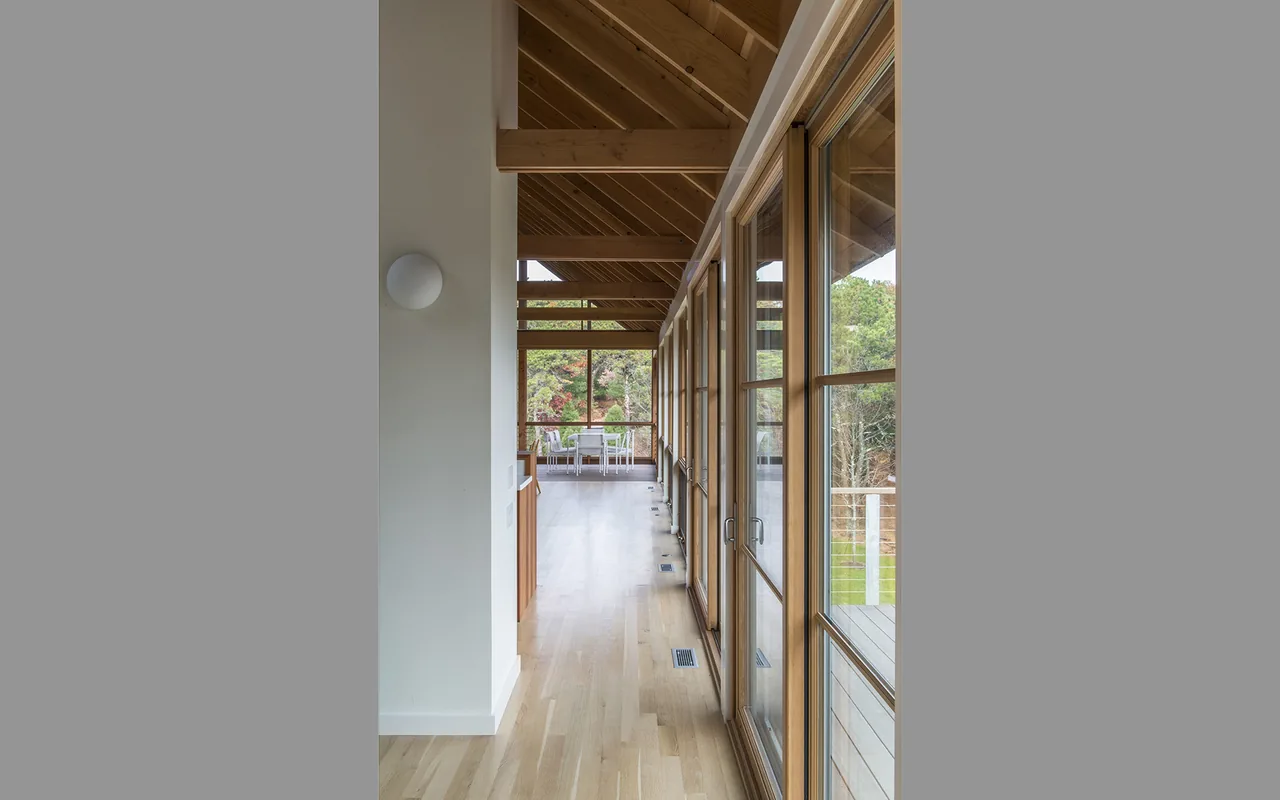 Long hallway with wooden truss ceiling and large windows looking out to a deck and wooded yard.