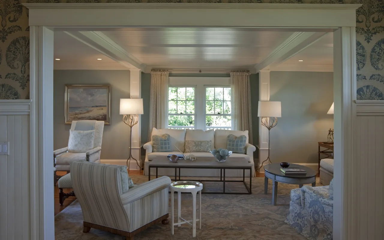 Traditional living room with central coffee table, cushioned armchairs, and natural light from double windows.