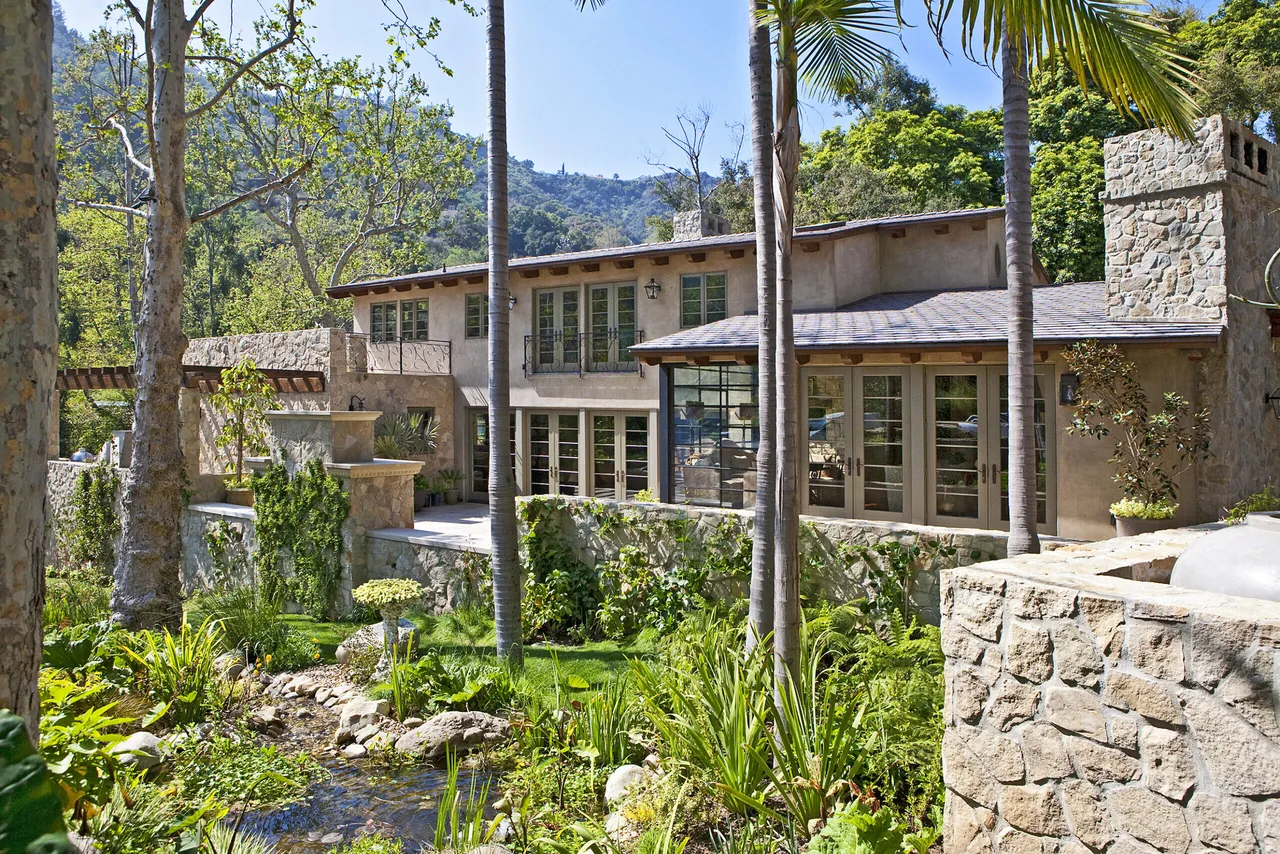 Two-story stone and stucco house with large windows and lush landscaping including palm trees and a small stream in the foreground.