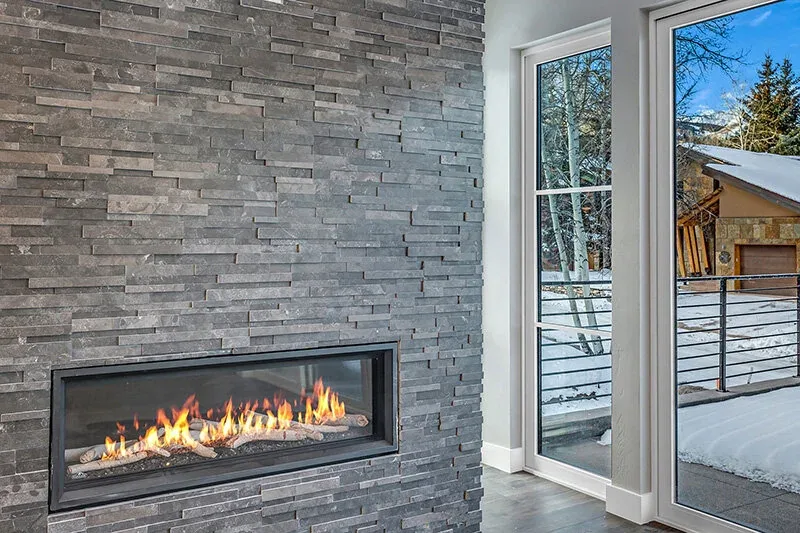 A dark stone wall houses a linear gas fireplace next to large vertical windows showing a snowy outdoor scene.