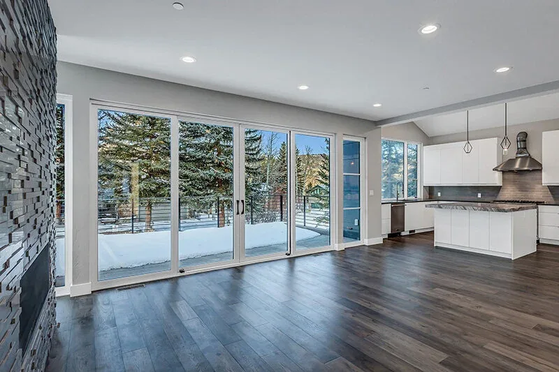 Living room and kitchen with large rectangular windows, dark wood flooring, and a dark stone fireplace showcasing a snowy outdoor view.
