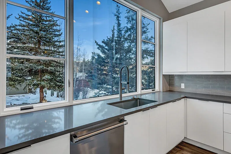A kitchen featuring white cabinets, gray countertops, a stainless steel dishwasher, and large windows with a snowy outdoor view.