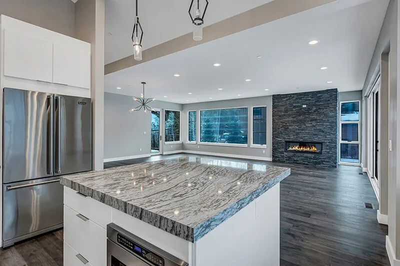 Open plan kitchen and living room feature large rectangular windows, dark wood flooring, and a dark stone fireplace with a metal frame.