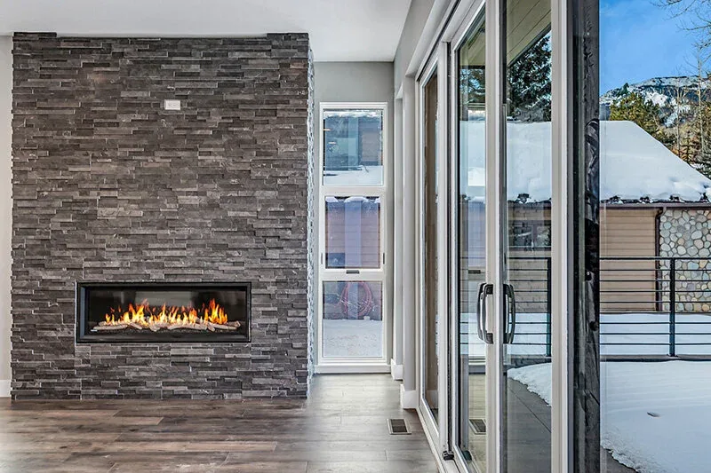 Dark stone fireplace with large sliding glass doors showing snow outside in the Snowmass Village Weekender project.