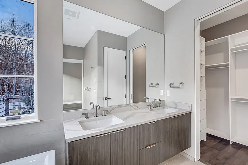 Bathroom vanity with double undermount sinks featuring a marbled countertop and faucet fixtures, adjacent to a walk-in closet and a window with a snowy view.