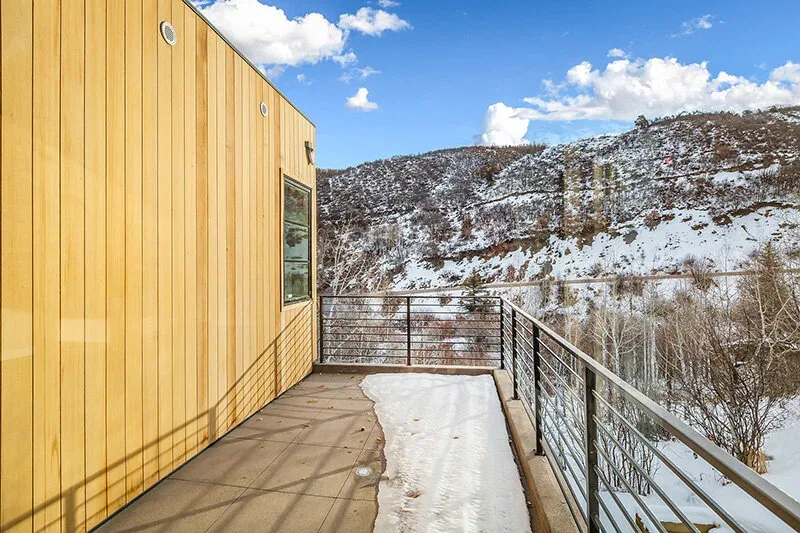 Balcony with vertical wood siding and metal cable railing facing a snow-covered hillside.