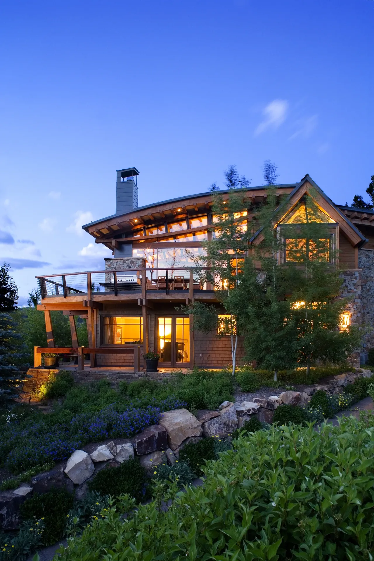 Twilight view of a two-story house with glowing interior lights, large deck, and stone landscaping.