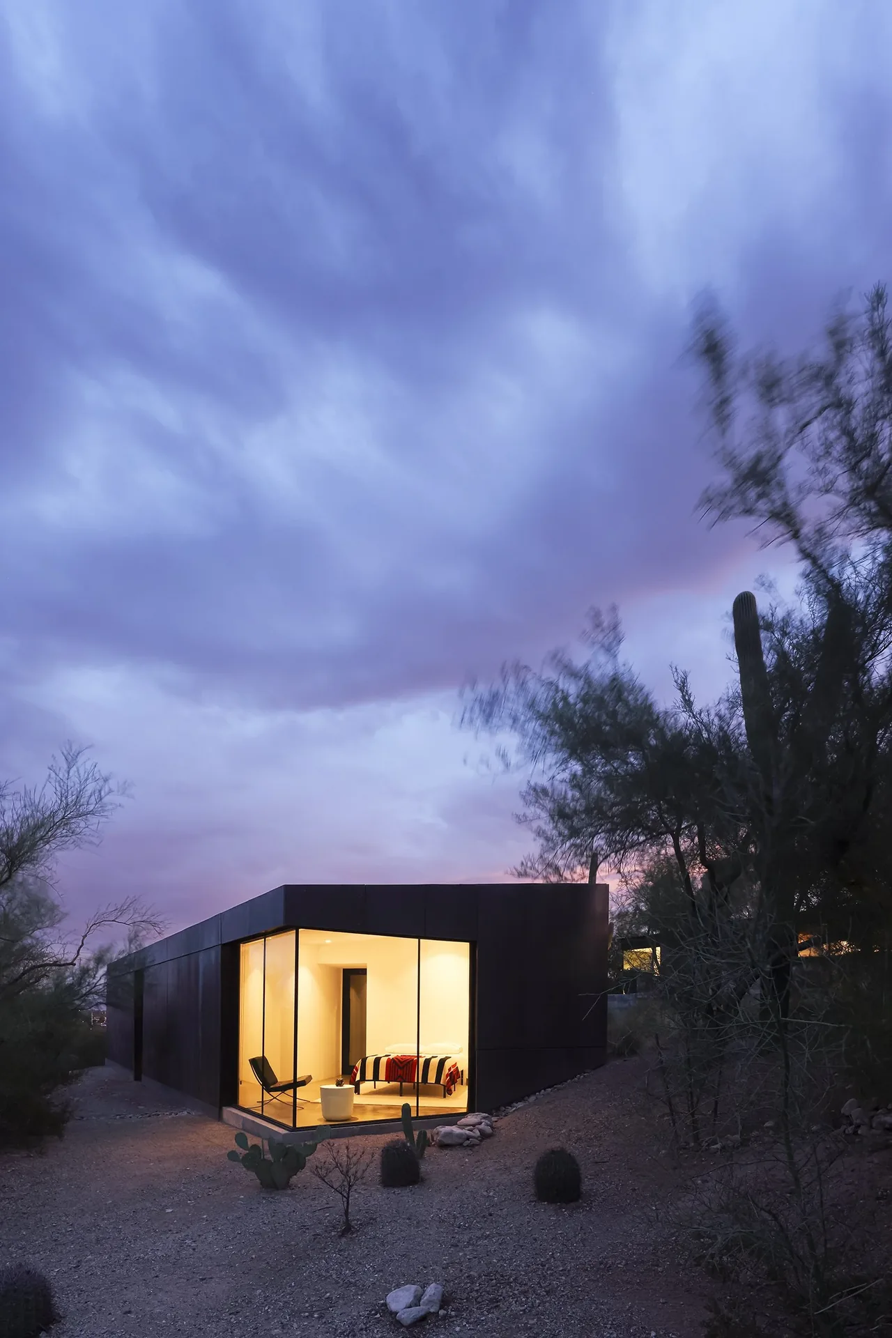 Casita OBSCURA features a blackened steel exterior with floor-to-ceiling corner windows revealing a softly lit bedroom interior.
