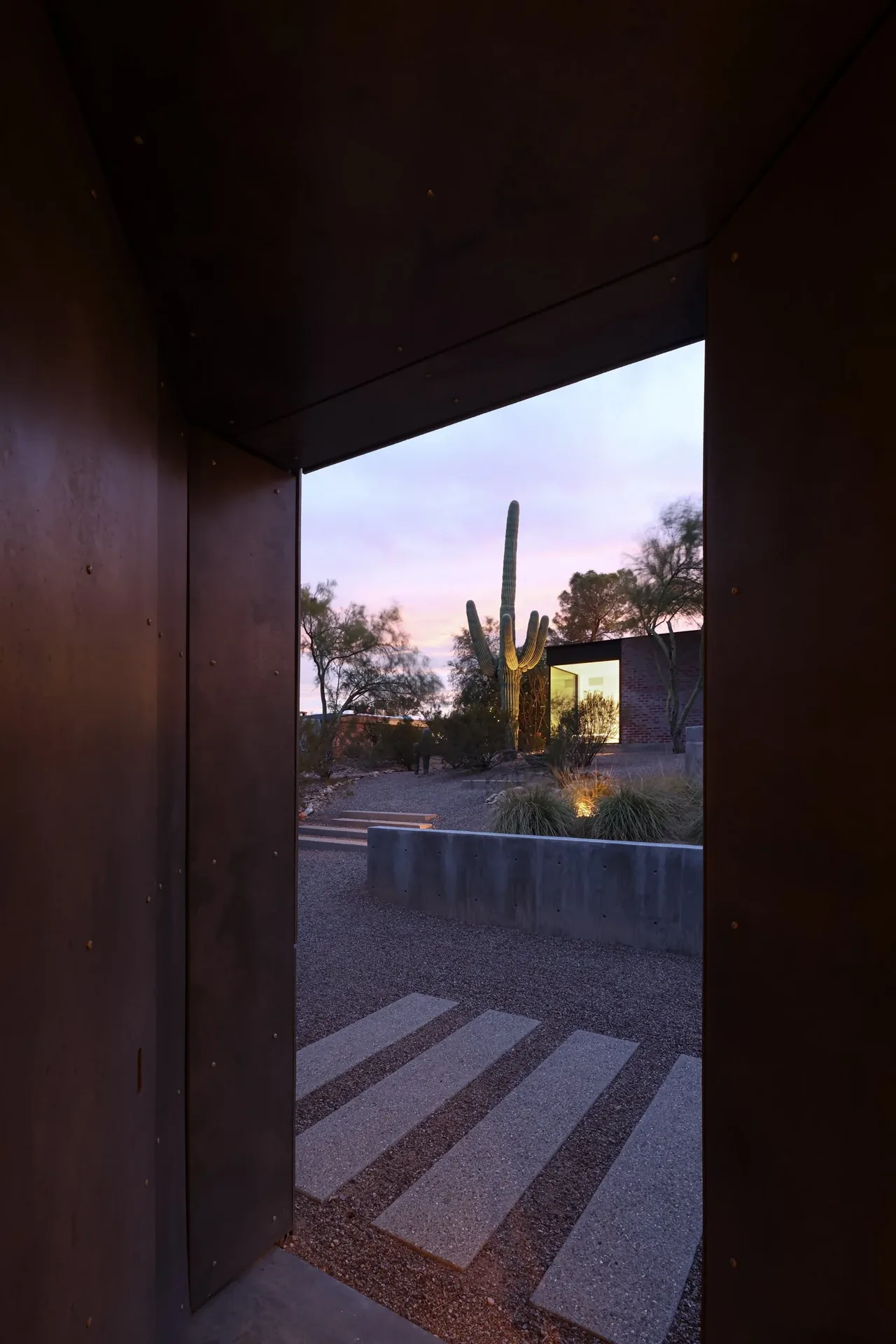 The casita OBSCURA project features a metal panel entrance framing desert landscaping and a rectangular volume building at dusk.
