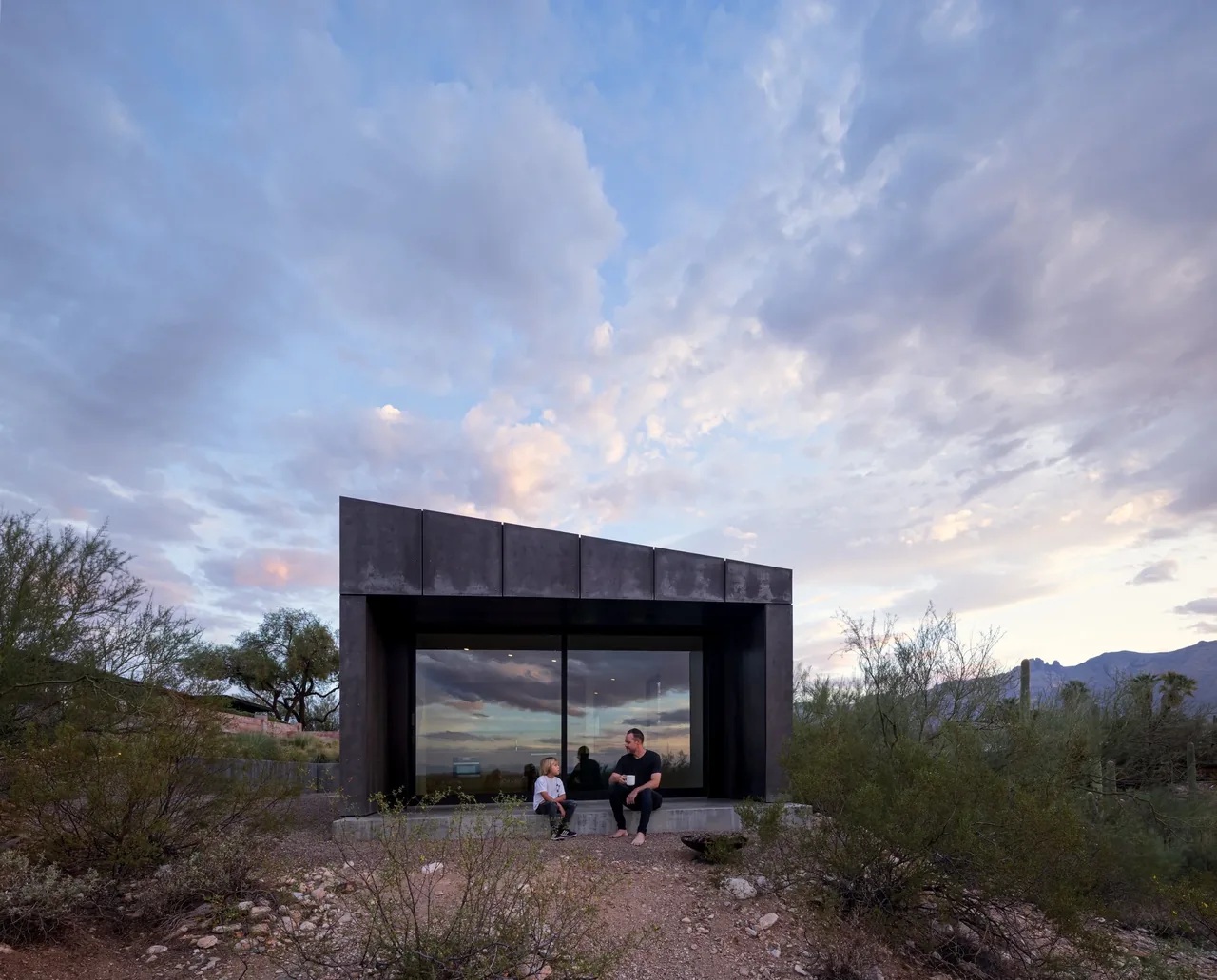 The casita OBSCURA features a black metal panel facade with floor-to-ceiling glazing, set in a desert landscape under a dramatic sky.