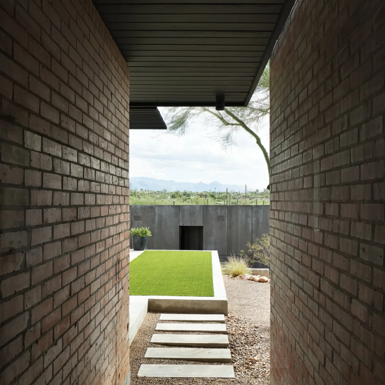 Narrow corridor between brick walls leads to a concrete patio with artificial turf and desert plants beyond.