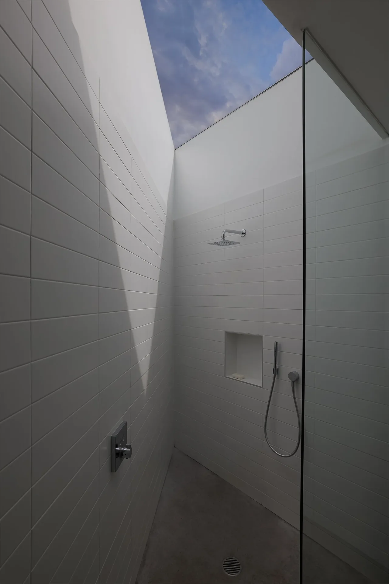 Outdoor shower with white horizontal tile walls and an open roof showing blue sky and clouds.