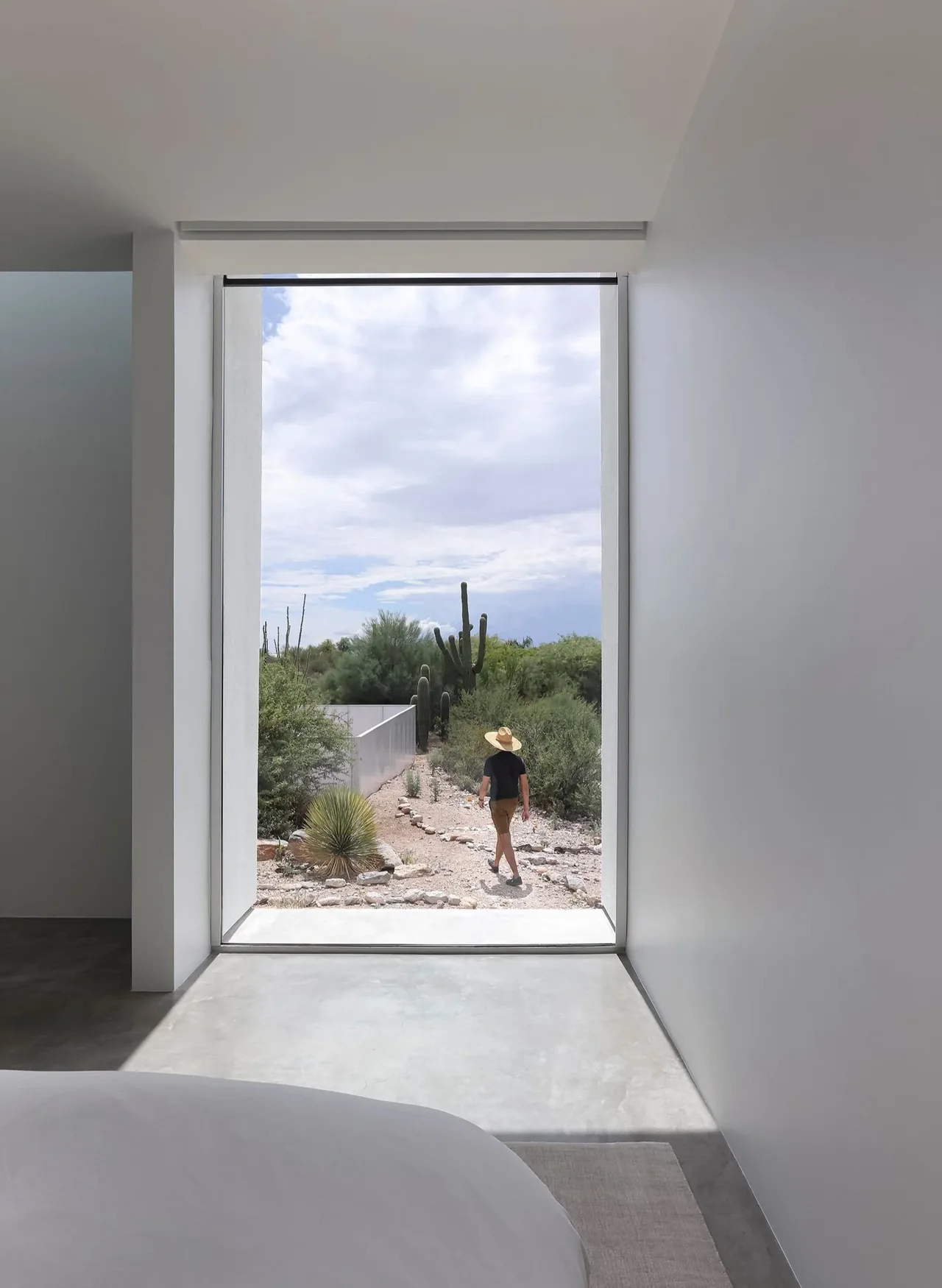 Minimal bedroom with floor-to-ceiling window framing a desert path and a person walking near tall cacti outside.