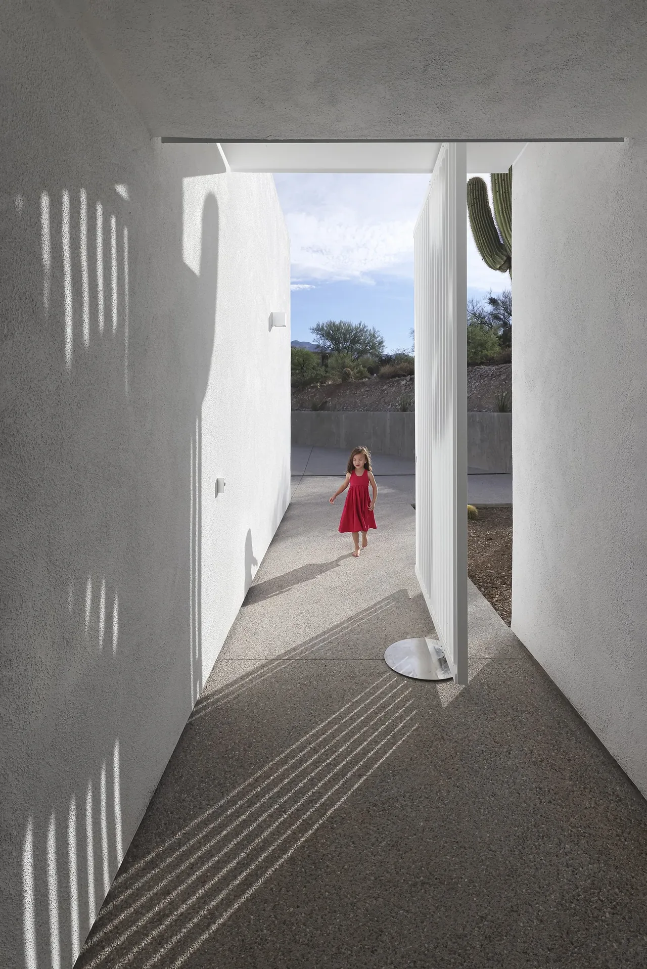 Narrow passageway with white textured walls and a pivoting vertical slat gate with desert landscape beyond.