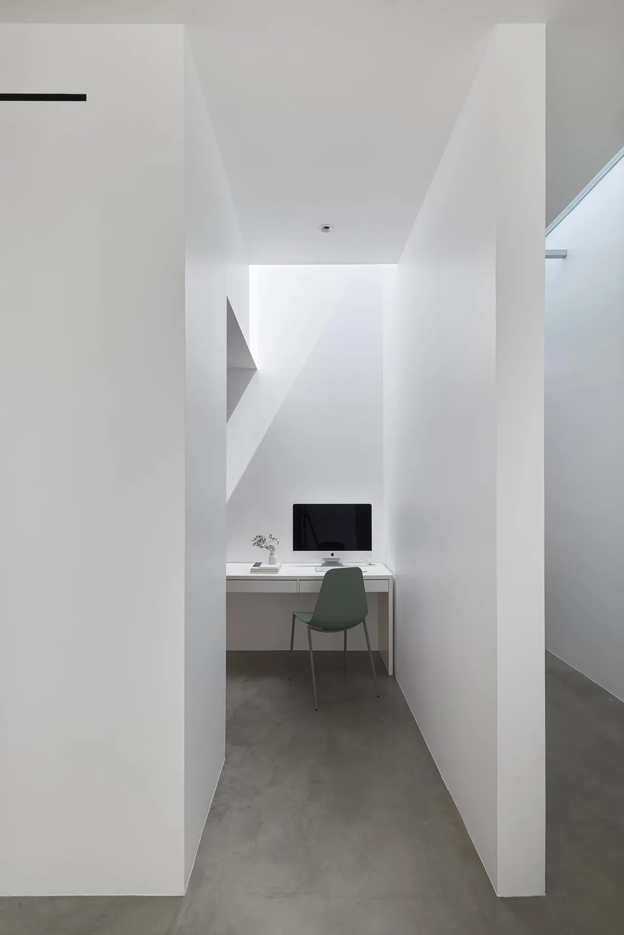 A minimalist office nook with a white desk, computer, green chair, and soft natural light from above.