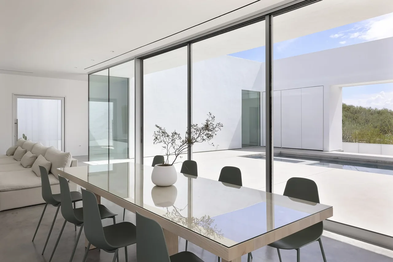 The dining space features a glass-top table, dark chairs, and expansive glazing overlooking a minimalist courtyard and pool.