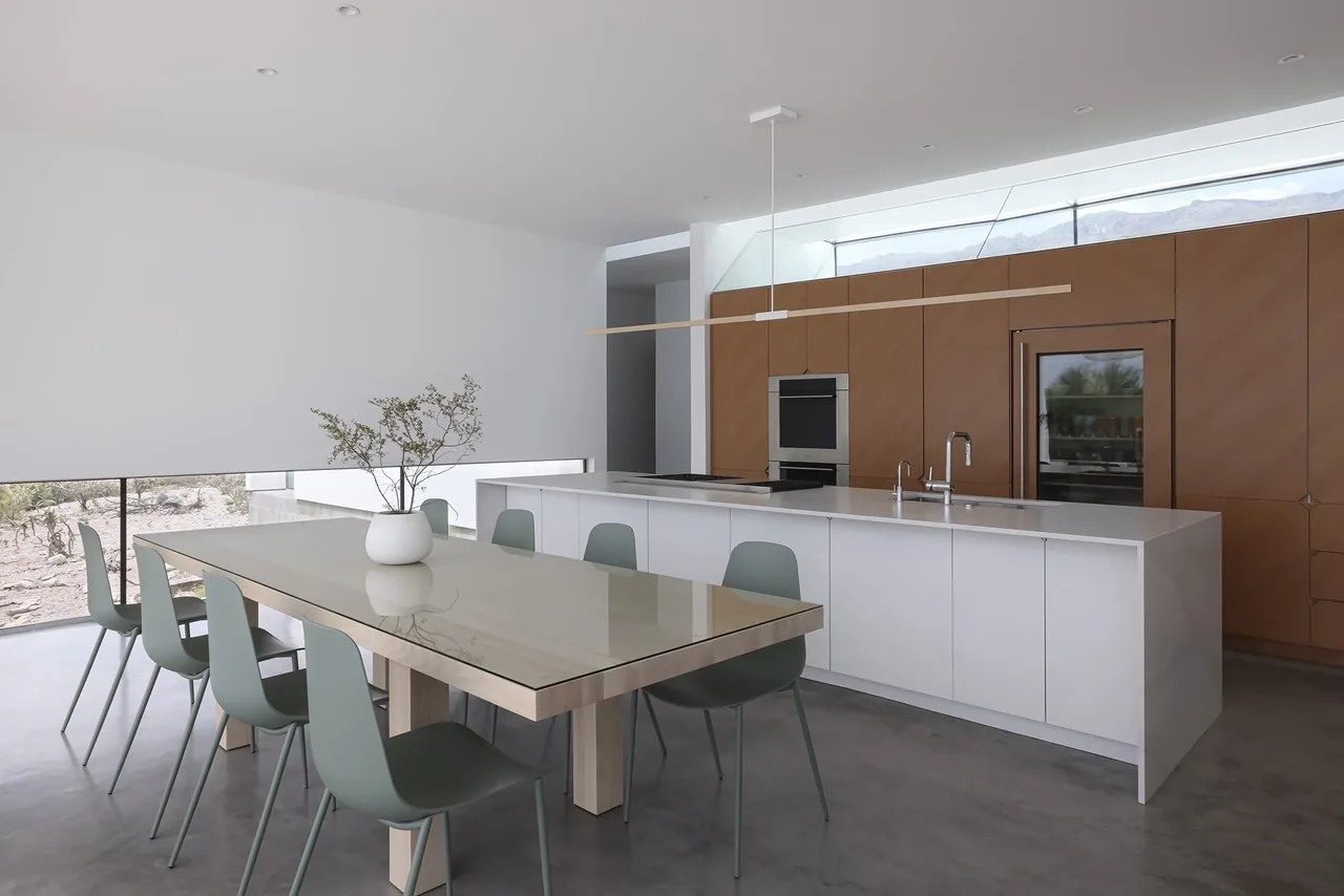 Kitchen and dining space with a white island, wood cabinetry, and a reflective glass dining table seating eight.