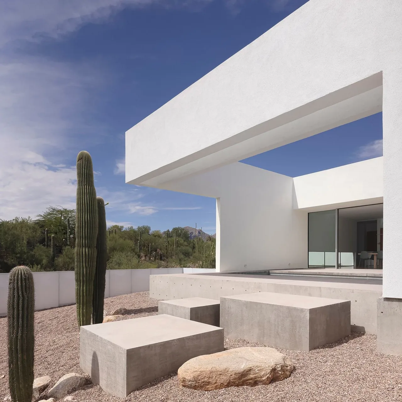 Exterior view of a modern white house with large rectangular concrete blocks in a gravel yard with tall cacti under a partly cloudy sky.