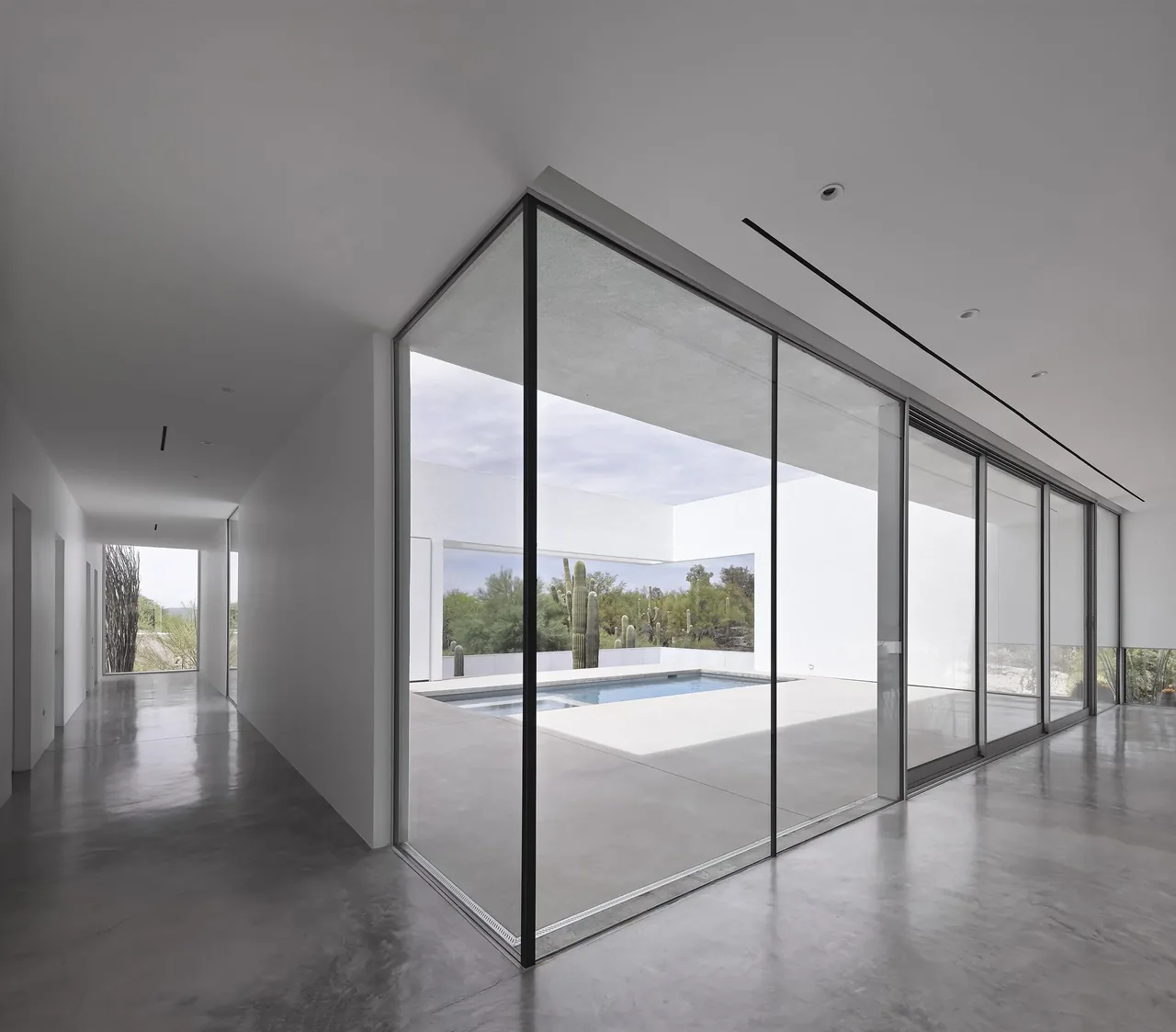 Minimalist interior hallway with polished concrete floor and large glass sliding doors looking onto a pool and desert landscaping with cacti.