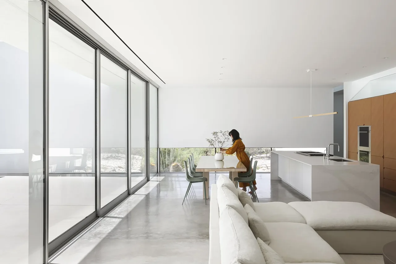 Open living and dining room with polished concrete floors and a large kitchen island, illuminated by natural light from floor-to-ceiling glazing.