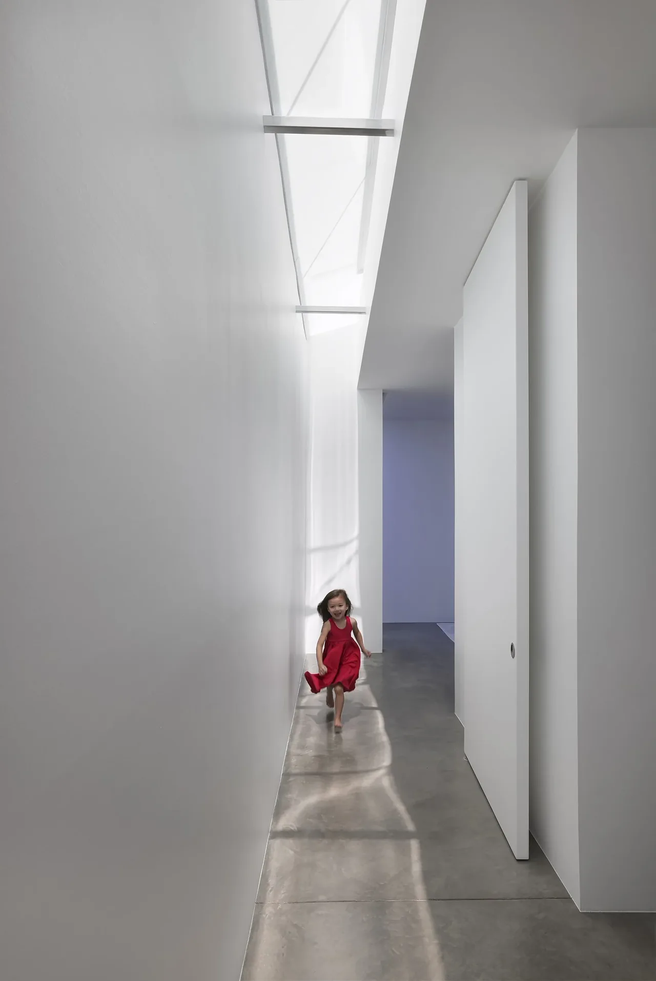 Narrow hallway with skylight illuminating a polished concrete floor where a child in a red dress runs.