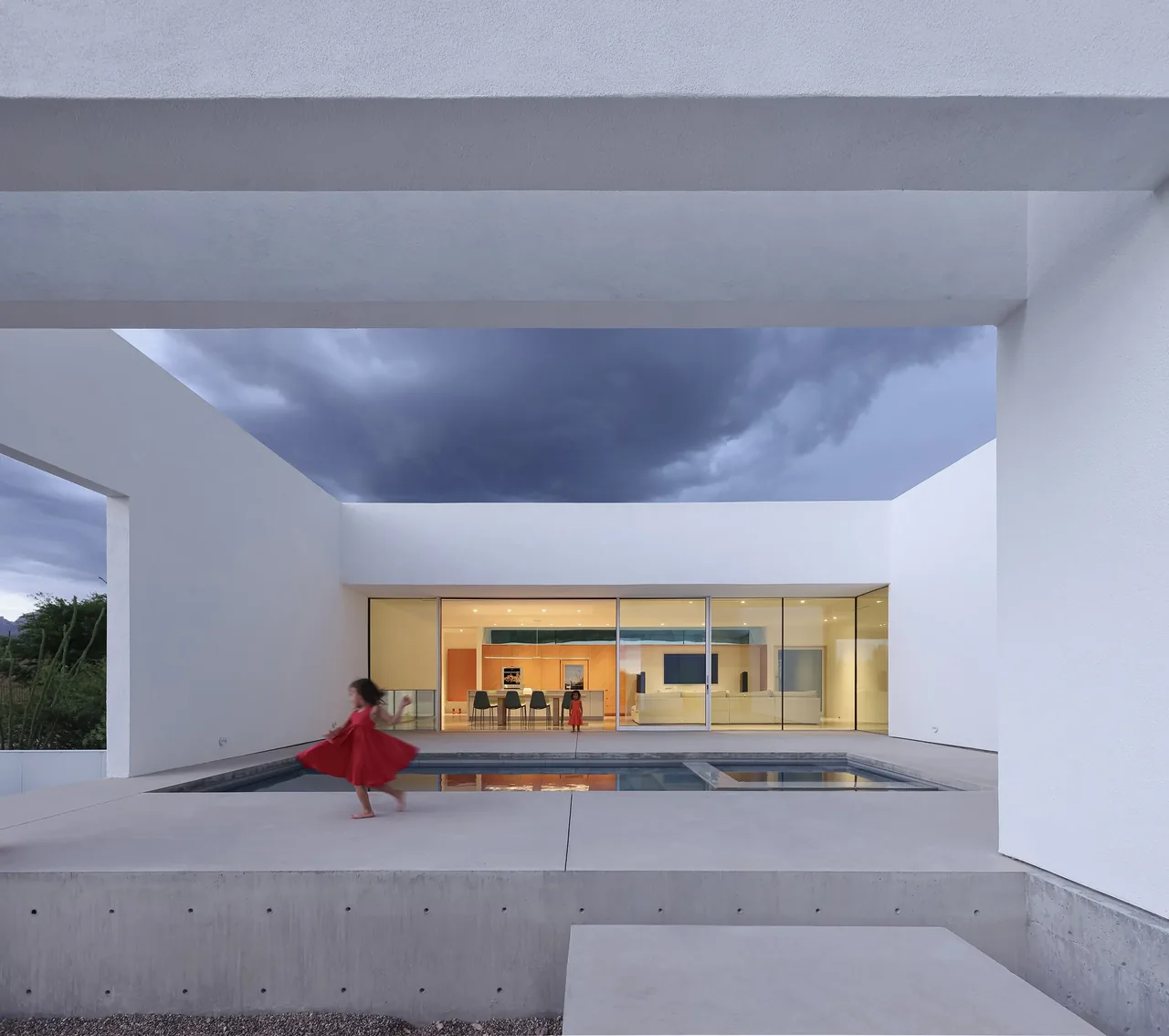 Exterior view of a modern white-walled house featuring large sliding glass doors and a minimalist pool under an overcast sky.