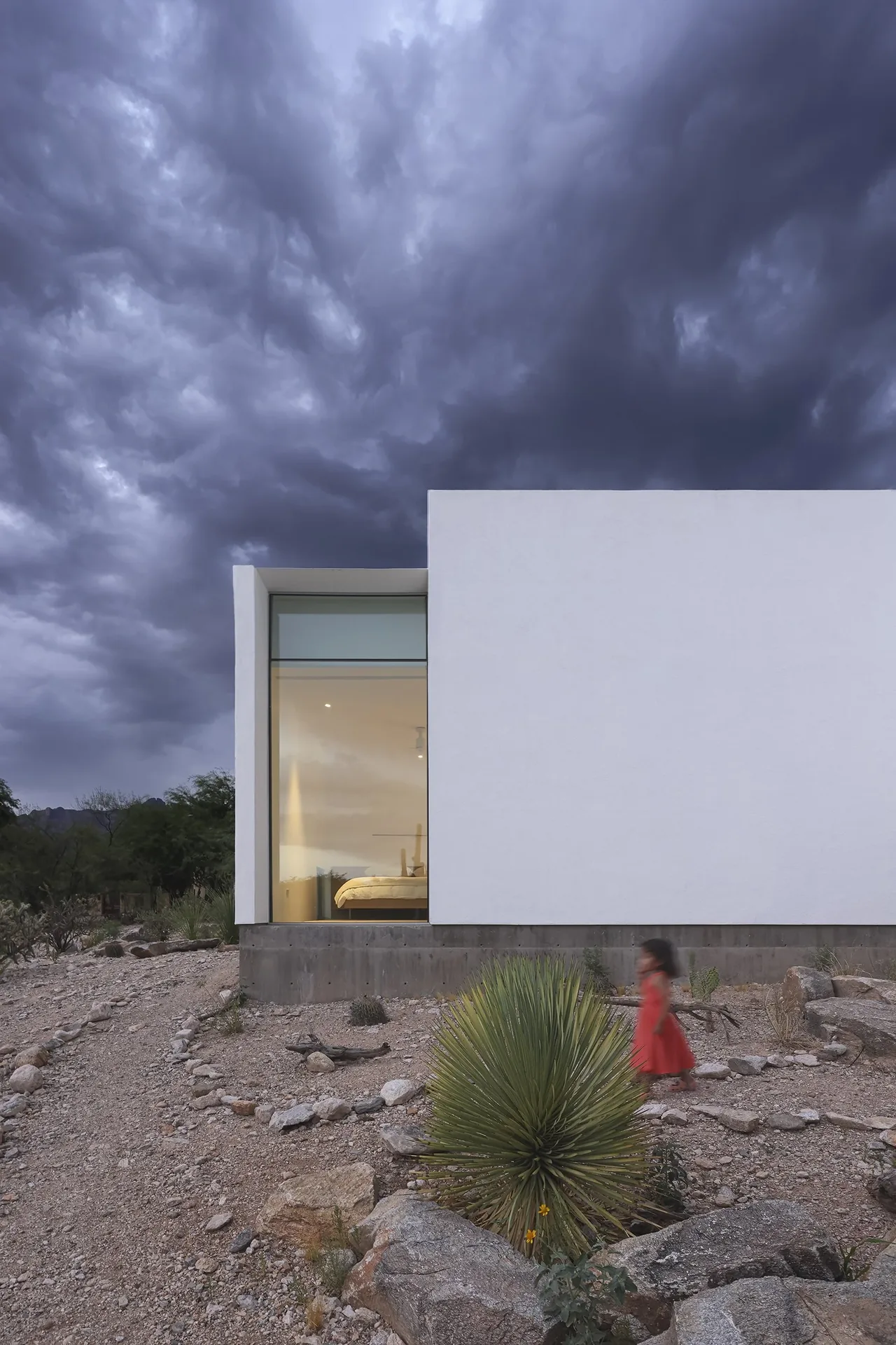 Exterior view of a white modern house with a large bedroom window and desert garden under stormy clouds.