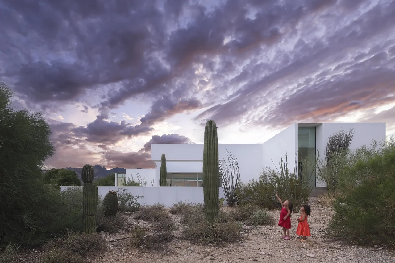 Modern white house with flat roof in desert landscape featuring cacti and dramatic sky at dusk.
