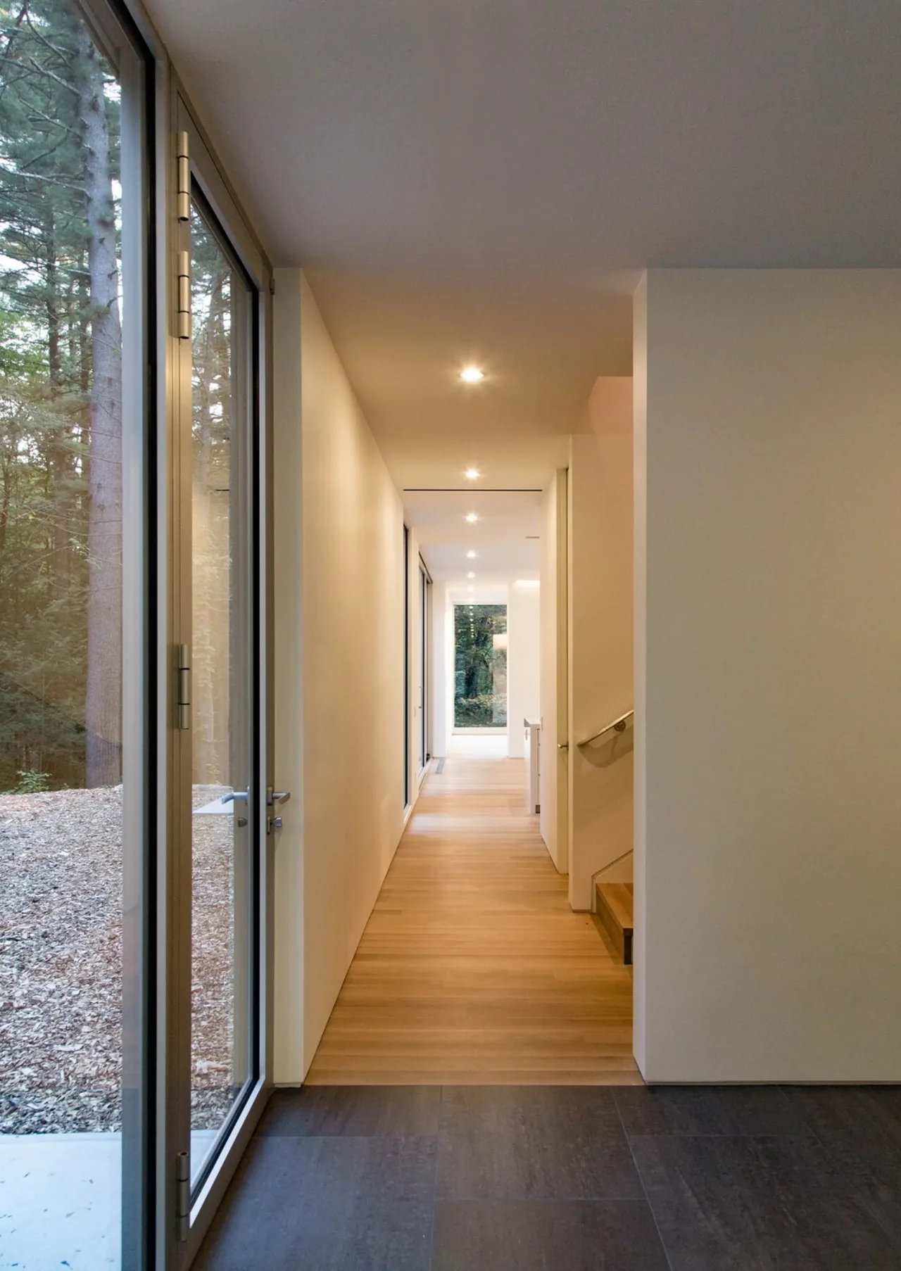 A hallway with wood flooring leads to a bright area with large windows at the end.