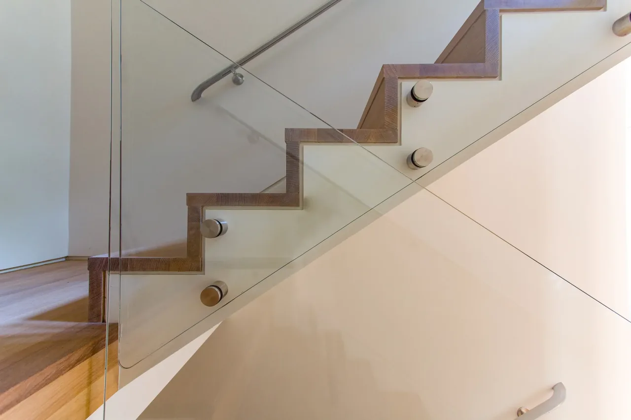 Interior view of a modern staircase with wooden treads and a glass balustrade supported by cylindrical metal fittings.