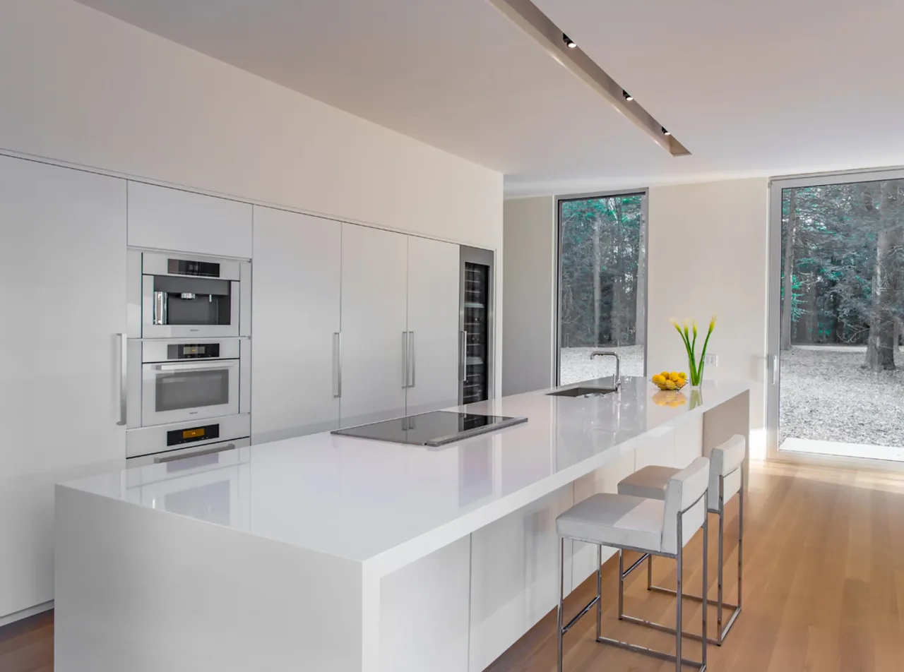 Kitchen in House No.207 by jacob lilley architects features a minimalist white island and large windows with views of the outdoors.