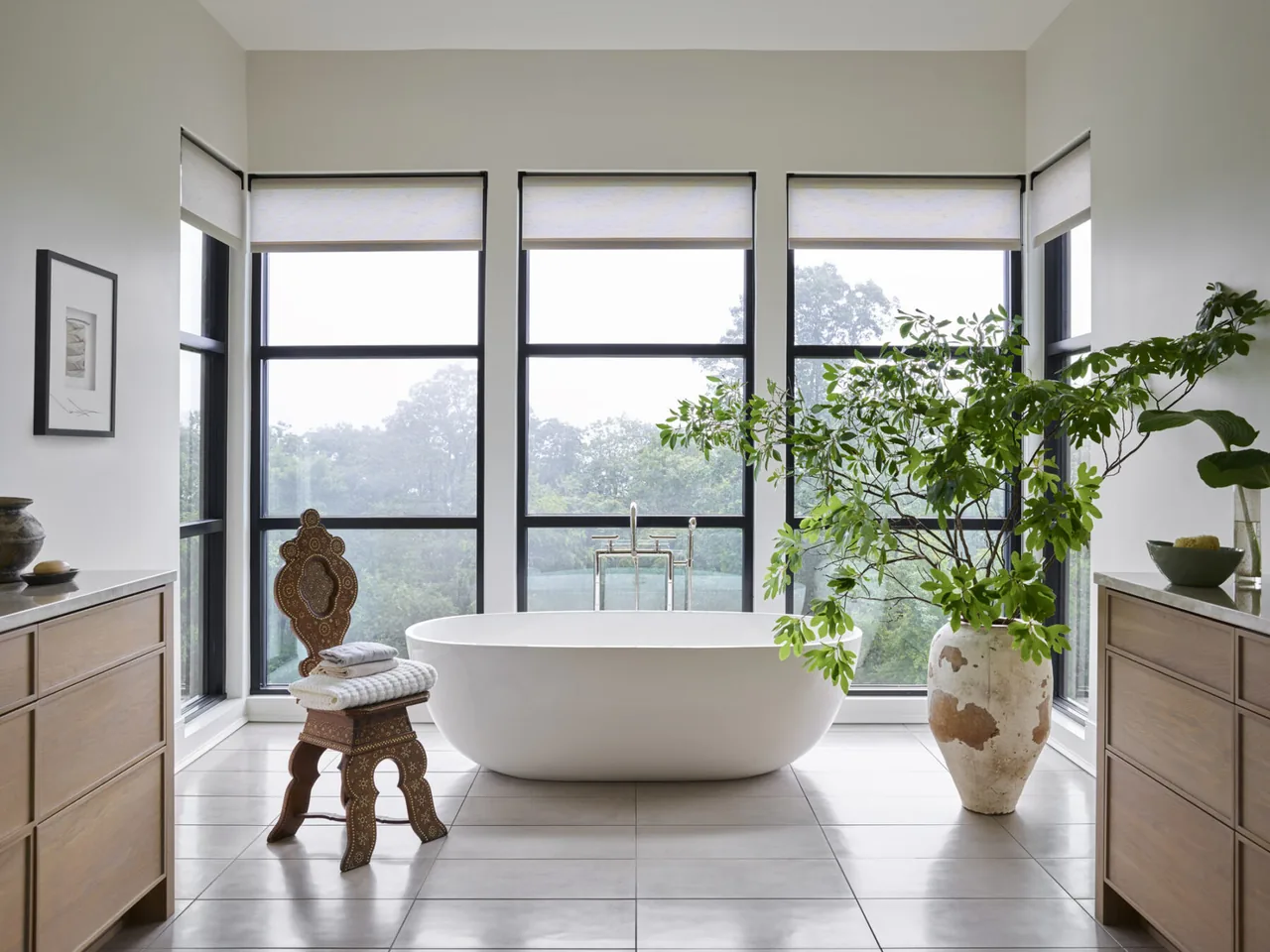 A bathroom features a white freestanding tub set in front of large black-framed windows with roller shades.