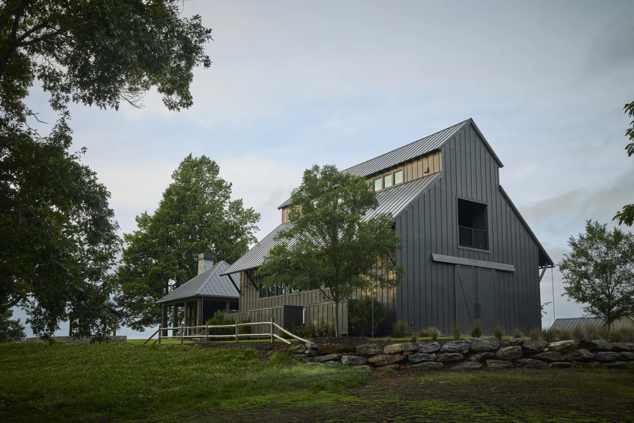 Modern barn-style house with standing seam metal siding, large windows, and surrounding landscaped rock wall and trees.