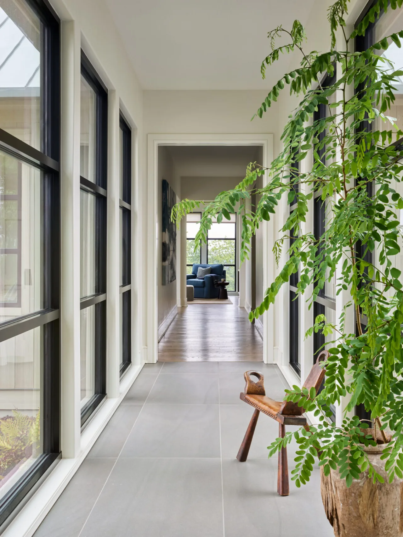 A bright hallway features large black-framed windows along both sides, a wooden bench, and a view into an adjacent sitting area.