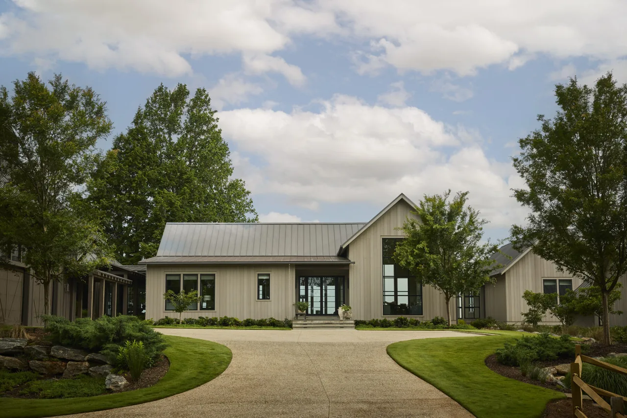 Single-story house with vertical siding and metal gable roof in landscaped setting with trees and curved driveway.