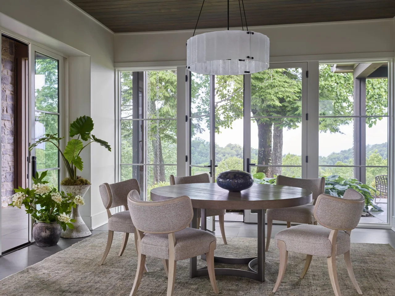 Dining room with round table and chairs illuminated by a modern circular chandelier with views of greenery through large glass doors.