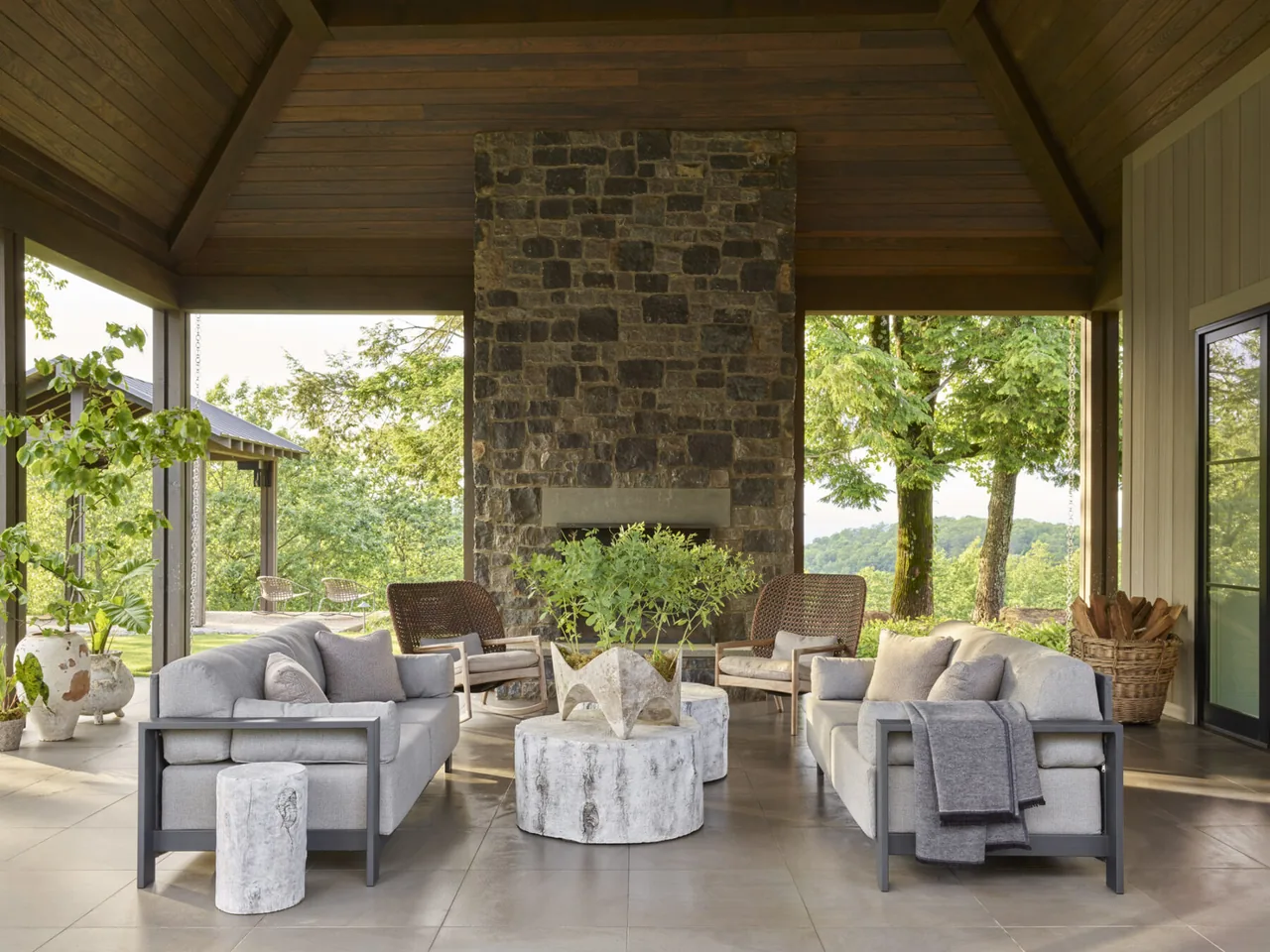 Covered patio with seating faces a tall stone fireplace, surrounded by large trees and landscape.