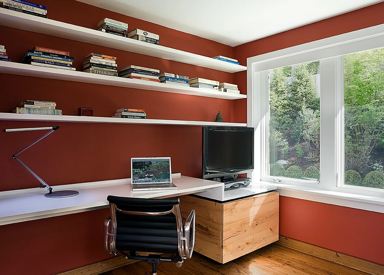A home office space with wood flooring, red walls, white shelves, and a corner desk with a laptop and TV.