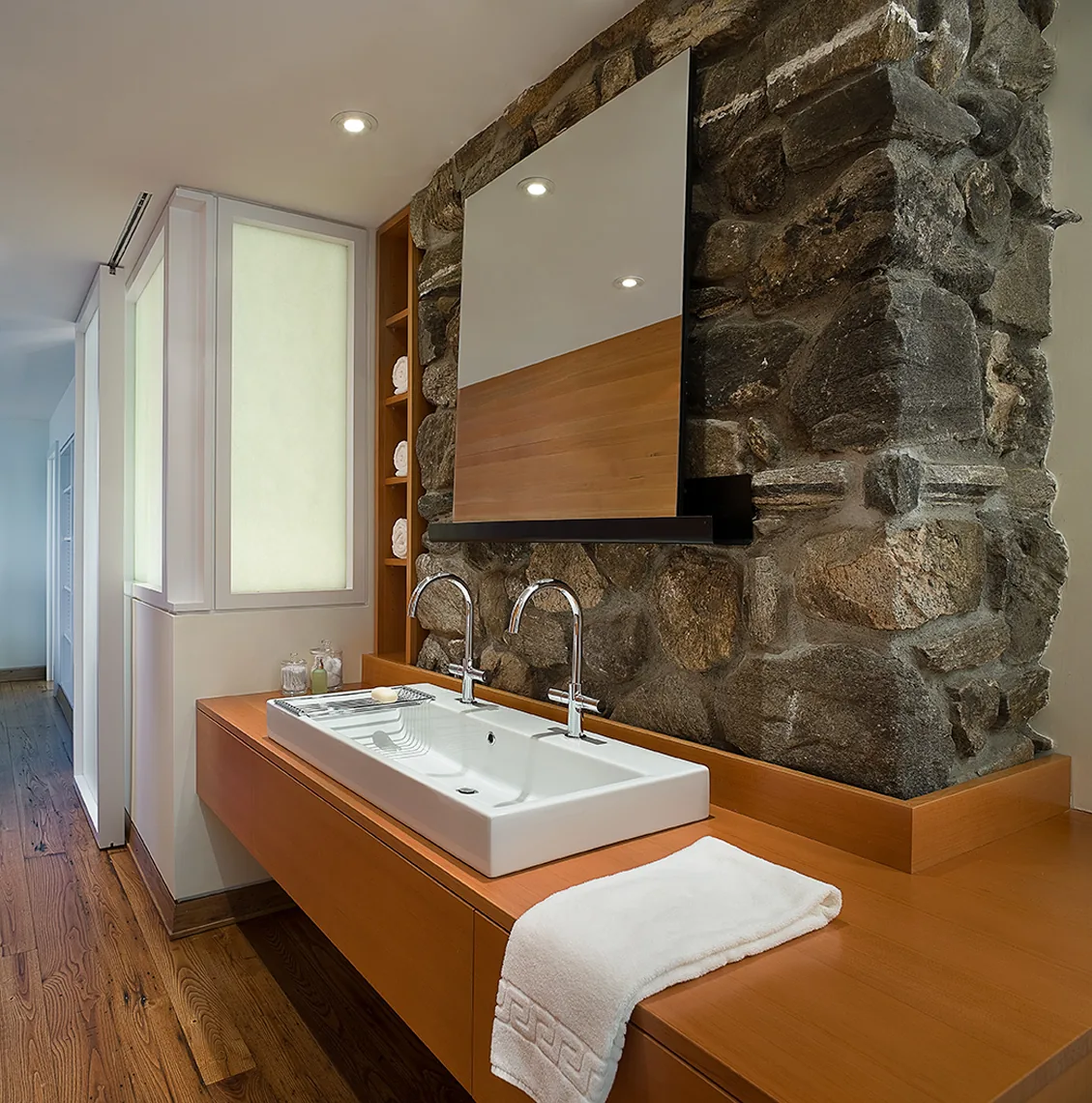 Bathroom sink with double faucets in a Connecticut lake house featuring a natural stone wall and wood cabinets.