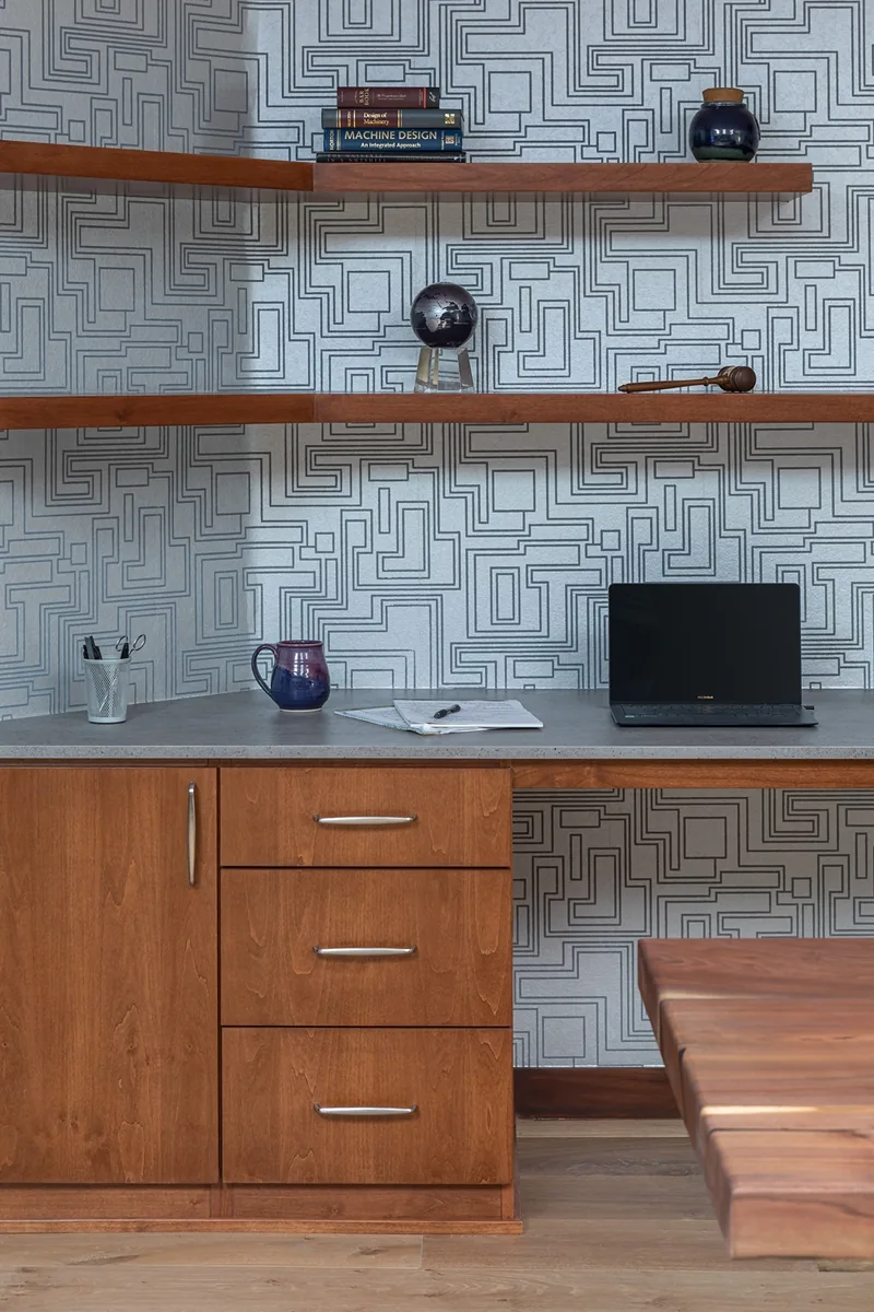 Home office desk with wooden cabinets and shelves, geometric wallpaper, and a laptop on the countertop.
