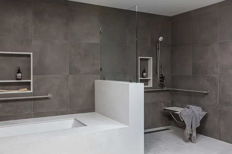 Bathroom featuring gray wall tiles, a built-in bathtub, and an accessible walk-in shower with a fold-down seat and stainless fixtures.